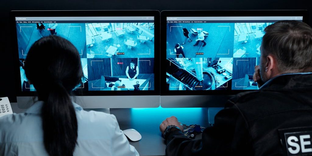 Rear view of male and female security guards sitting in front of desktop computers with video camera on screens in surveillance room